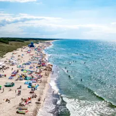 Tłum ludzi na plaży w Darłowie, korzystających z uroków Bałtyku. Wiele kolorowych parawanów i namiotów, a w wodzie kąpiący się. To typowe atrakcje Darłowa latem.