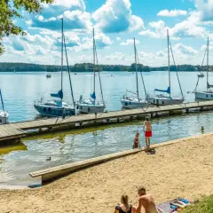 Słoneczny dzień na mazurskich jeziorach. Cumujące jachty, piaszczysta plaża i ludzie odpoczywający nad wodą.
