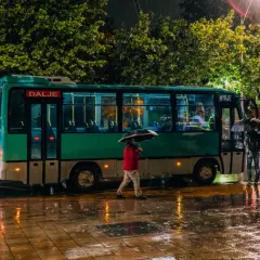 Deszczowy wieczór na lotnisku Tirana. Ludzie z parasolami czekają na autobus, a mokry chodnik odbija światła.