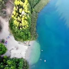 Plaża nad jeziorem tętni życiem. Ludzie odpoczywają na piasku, kąpią się w krystalicznie czystej wodzie, otoczeni zielenią drzew.