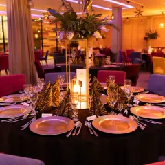 Elegancko nakryte stoły w restauracji, gotowe na przyjęcie gości. W centralnym punkcie stołu znajduje się ozdobna kompozycja z liści i świeca, tworząc ciepłą atmosferę. Kolorowe krzesła dodają wnętrzu charakteru. To idealne miejsce na wykwintne jedzeni...