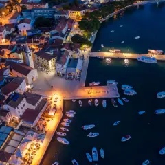 Nadmorskie miasteczko nocą, z oświetlonymi domami i portem pełnym łodzi. W oddali widać fazana plaże.