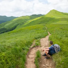 Turysta odpoczywa na szlaku, podziwiając zielone połoniny. Gdzie w Bieszczady? Tu, gdzie ścieżka wije się wśród traw ku szczytom.