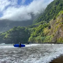 Surfer z deską na plaży na Maderze, bez samochodu. W tle góry porośnięte zielenią i chmury.