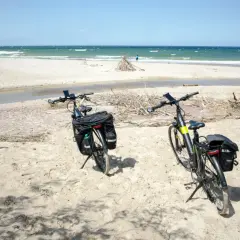 Dwa rowery z sakwami na piaszczystej plaży, gotowe na odkrywanie tras rowerowych nad morzem. W tle widać morze i niebo.