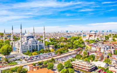 Widok na Stambuł, miasto łączące Europę i Azję, z Błękitnym Meczetem i Hagia Sophia. W tle nowoczesne wieżowce, a na pierwszym planie zielone drzewa i zabudowa miejska.