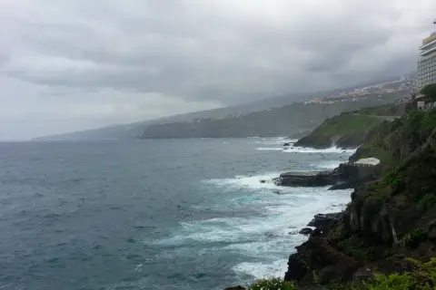 Mroczne chmury nad oceanem. Teneryfa pogoda w styczniu, fale rozbijają się o skaliste wybrzeże, w oddali widać hotel.