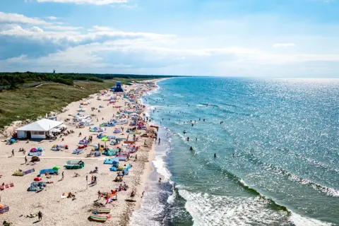 Tłum ludzi na plaży w Darłowie, korzystających z uroków Bałtyku. Wiele kolorowych parawanów i namiotów, a w wodzie kąpiący się. To typowe atrakcje Darłowa latem.