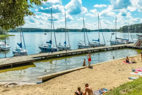 Słoneczny dzień na mazurskich jeziorach. Cumujące jachty, piaszczysta plaża i ludzie odpoczywający nad wodą.