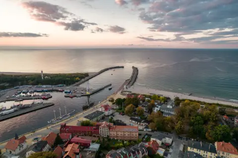 Widok z lotu ptaka na port i plażę w Ustce. Widać liczne łodzie, latarnię morską i falochrony. To idealne miejsce na odkrywanie ustka atrakcje.