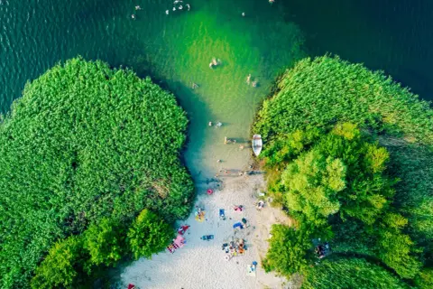 Letnie, magiczne jeziora w Polsce. Ludzie kąpią się w zielonej wodzie, odpoczywają na piaszczystej plaży otoczonej trzcinami i drzewami.