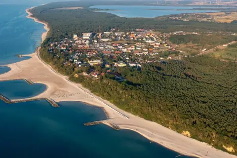 Jarosławiec: widok z lotu ptaka na plażę z falochronami, las i zabudowania.