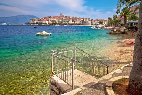 Widok na turkusowe morze i kamienistą plażę z kamiennymi schodami prowadzącymi do wody. W oddali widać urokliwe miasto Korcula, pełne zabytków i atrakcji.