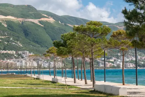 Wlora, Albania: nadmorska promenada z sosnami, turkusowe morze i góry w tle.