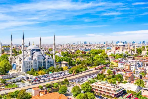 Widok na Stambuł, miasto łączące Europę i Azję, z Błękitnym Meczetem i Hagia Sophia. W tle nowoczesne wieżowce, a na pierwszym planie zielone drzewa i zabudowa miejska.