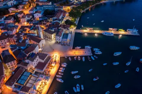 Nadmorskie miasteczko nocą, z oświetlonymi domami i portem pełnym łodzi. W oddali widać fazana plaże.