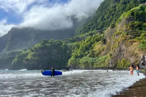 Surfer z deską na plaży na Maderze, bez samochodu. W tle góry porośnięte zielenią i chmury.