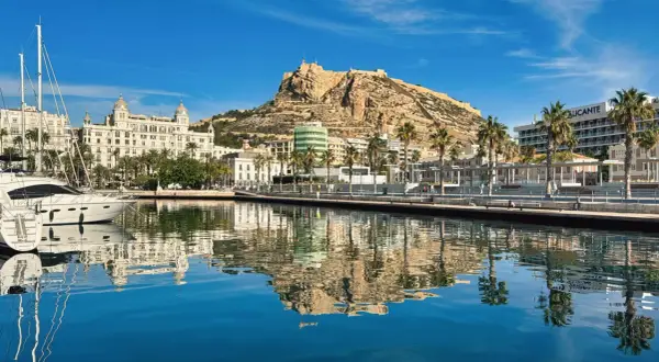 Widok na port i zamek Santa Barbara w Alicante. Luksusowe jachty odbijają się w spokojnej wodzie.