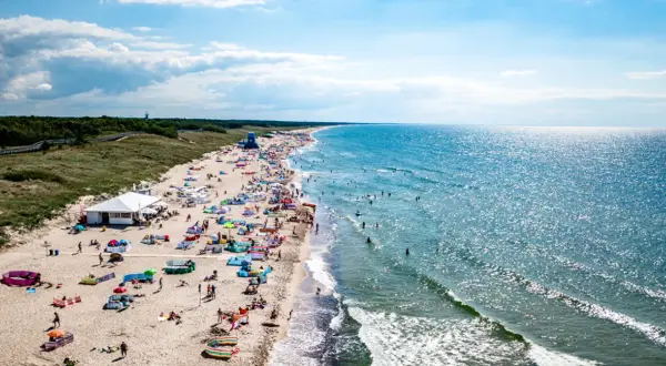 Tłum ludzi na plaży w Darłowie, korzystających z uroków Bałtyku. Wiele kolorowych parawanów i namiotów, a w wodzie kąpiący się. To typowe atrakcje Darłowa latem.