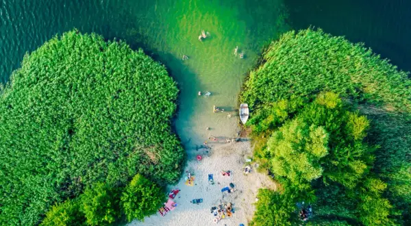 Letnie, magiczne jeziora w Polsce. Ludzie kąpią się w zielonej wodzie, odpoczywają na piaszczystej plaży otoczonej trzcinami i drzewami.