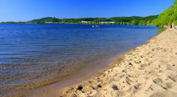 Słoneczny dzień nad zalewem Sulejowskim. Piaszczysta plaża, błękitna woda i zielone wzgórza w tle.