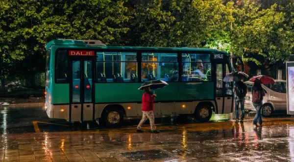Deszczowy wieczór na lotnisku Tirana. Ludzie z parasolami czekają na autobus, a mokry chodnik odbija światła.