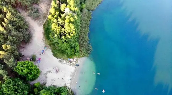 Plaża nad jeziorem tętni życiem. Ludzie odpoczywają na piasku, kąpią się w krystalicznie czystej wodzie, otoczeni zielenią drzew.