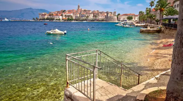 Widok na turkusowe morze i kamienistą plażę z kamiennymi schodami prowadzącymi do wody. W oddali widać urokliwe miasto Korcula, pełne zabytków i atrakcji.