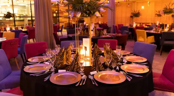 Elegancko nakryte stoły w restauracji, gotowe na przyjęcie gości. W centralnym punkcie stołu znajduje się ozdobna kompozycja z liści i świeca, tworząc ciepłą atmosferę. Kolorowe krzesła dodają wnętrzu charakteru. To idealne miejsce na wykwintne jedzeni...