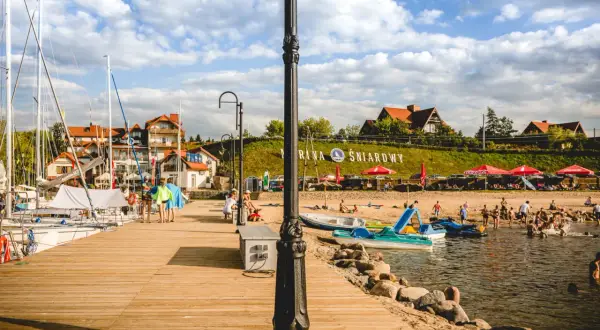 Letnia przygoda w krainie tysiąca jezior. Drewniane molo, żaglówki, plaża z ludźmi i budynki nad jeziorem.