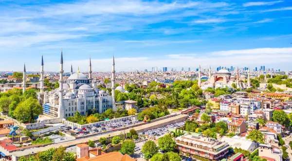 Widok na Stambuł, miasto łączące Europę i Azję, z Błękitnym Meczetem i Hagia Sophia. W tle nowoczesne wieżowce, a na pierwszym planie zielone drzewa i zabudowa miejska.