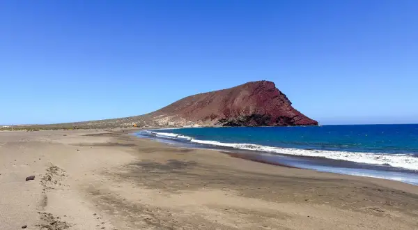 Pusta plaża na Teneryfie z charakterystyczną czerwoną górą w tle i turkusowym oceanem. Idealne miejsce na relaks.