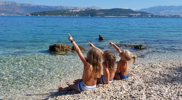 Trzy dziewczynki siedzą na kamienistej plaży w Chorwacji, wskazując palcami na morze i góry. To idealne chorwackie atrakcje dla dzieci.