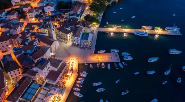Nadmorskie miasteczko nocą, z oświetlonymi domami i portem pełnym łodzi. W oddali widać fazana plaże.