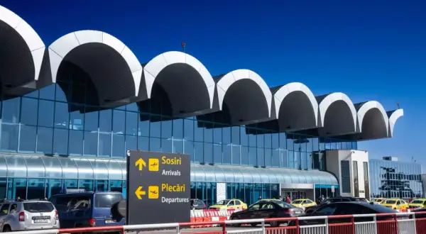 Nowoczesny terminal lotniska w Bukareszcie z charakterystycznymi łukami dachu. Tablice kierują do przylotów i odlotów.