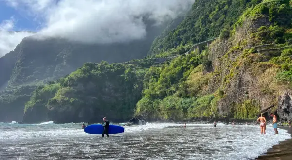 Surfer z deską na plaży na Maderze, bez samochodu. W tle góry porośnięte zielenią i chmury.