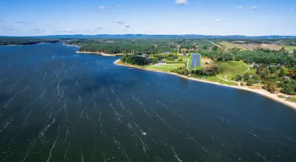 Malownicze jezioro Hańcza z piaszczystą plażą i lasem. W oddali widać farmę fotowoltaiczną, która jest jedną z atrakcji regionu.
