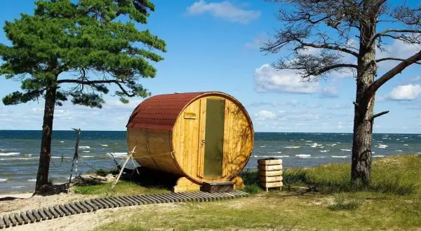 Beczka-sauna nad morzem, jedna z nietypowych atrakcji Juraty. Drewniana konstrukcja z czerwonym dachem, przy plaży, pod drzewami.