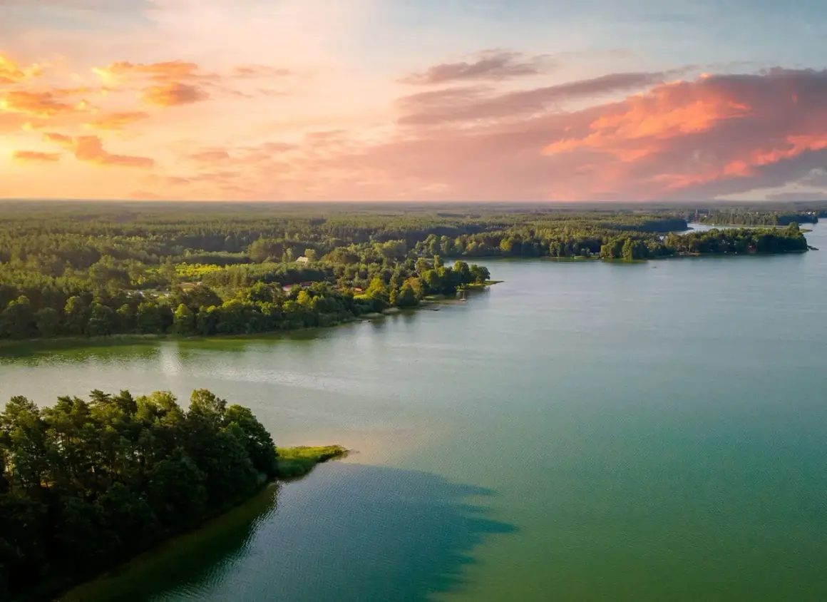 Zachód słońca nad jeziorem, z lasem i domkami w tle. Idealne, fajne miejsca nad jeziorem na relaks.