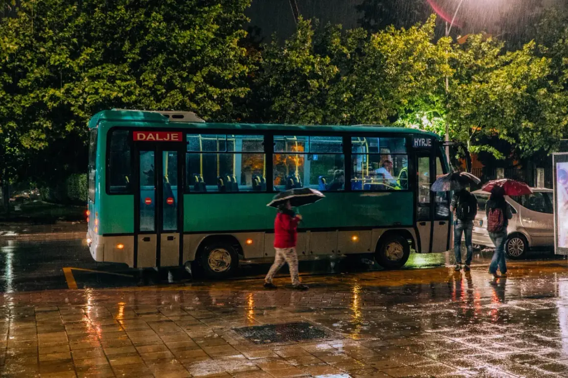 Deszczowy wieczór na lotnisku Tirana. Ludzie z parasolami czekają na autobus, a mokry chodnik odbija światła.