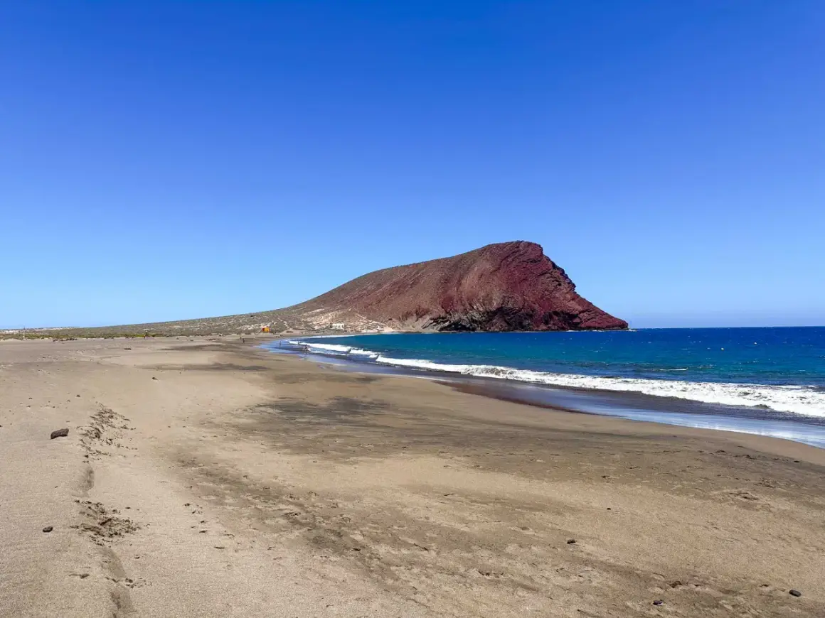 Pusta plaża na Teneryfie z charakterystyczną czerwoną górą w tle i turkusowym oceanem. Idealne miejsce na relaks.