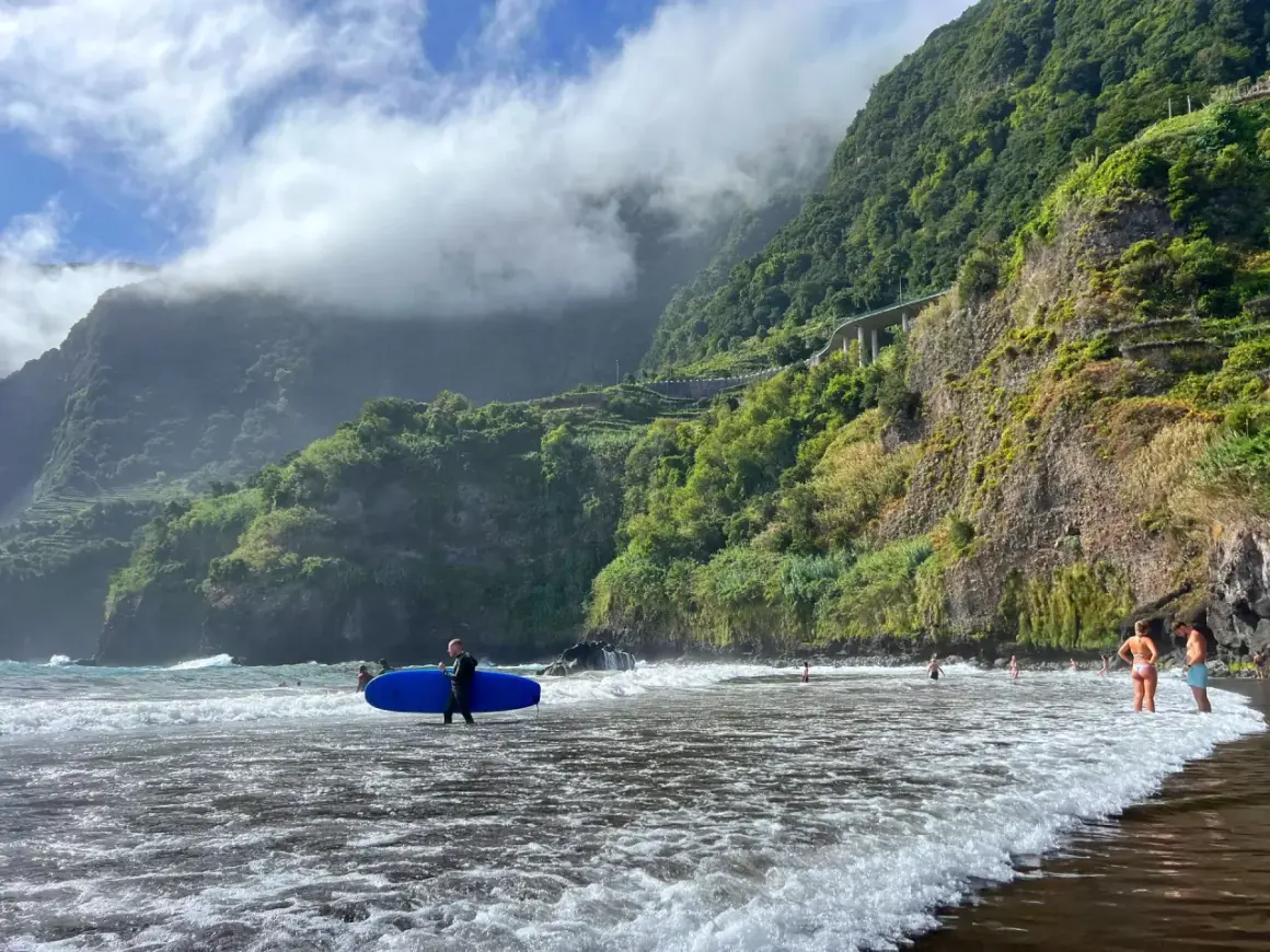 Surfer z deską na plaży na Maderze, bez samochodu. W tle góry porośnięte zielenią i chmury.