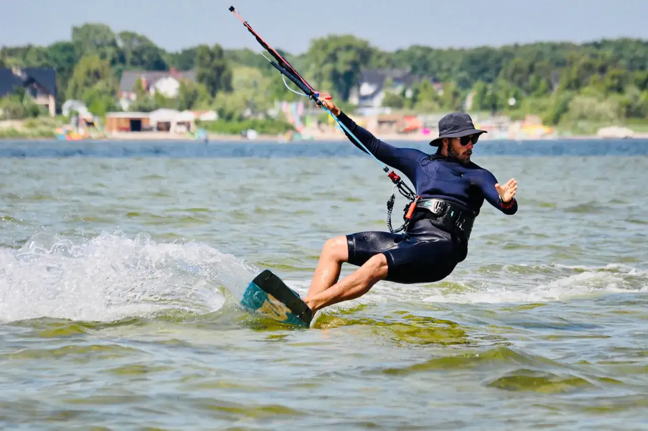 Mężczyzna w piance i kapeluszu uprawia kitesurfing w Polsce, tworząc pianę na wodzie.