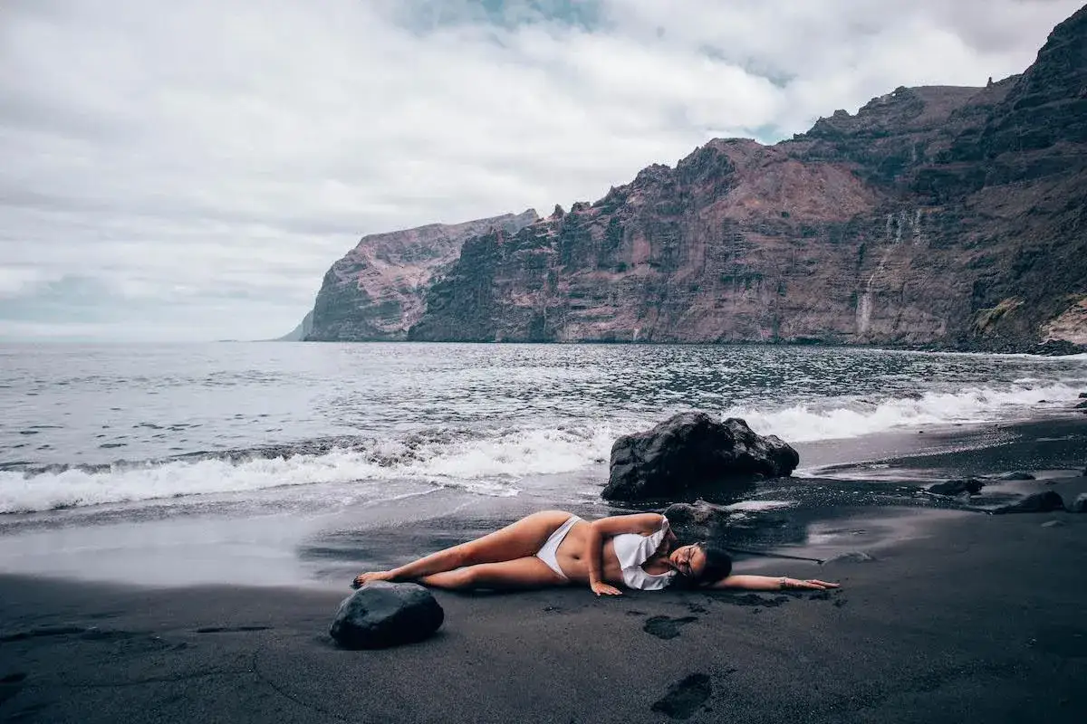 Kobieta w białym bikini leży na czarnej plaży na Teneryfie południe, z falami obmywającymi brzeg i skalistymi klifami w tle.