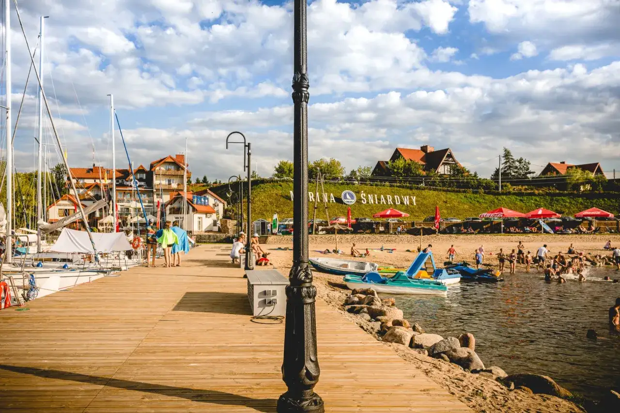Słoneczny dzień nad jeziorem Śniardwy. Drewniane molo, żagl&oacute;wki, plaża z ludźmi i charakterystyczny napis 