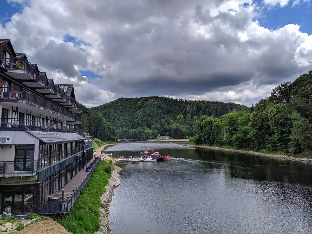 Hotel nad jeziorem Bystrzyckim, z tarasami i widokiem na zalesione wzg&oacute;rza. Na wodzie widać ł&oacute;dki.