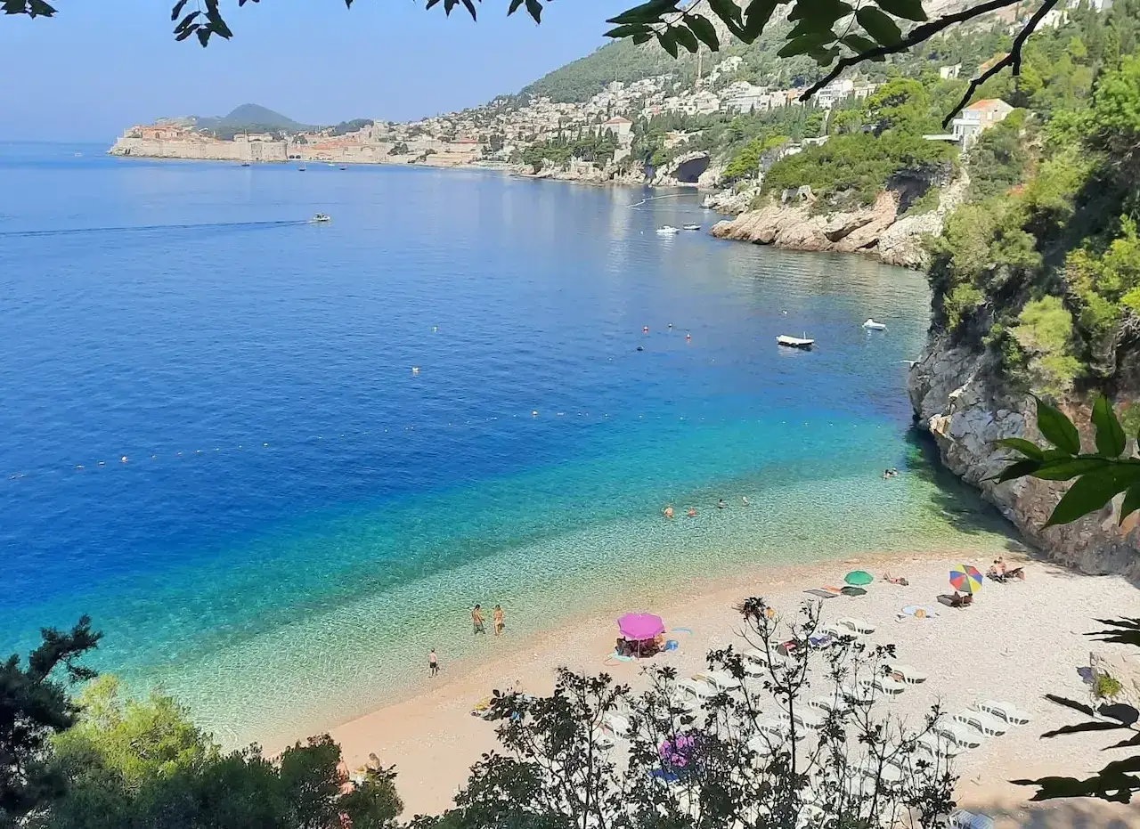 Malownicza zatoka z turkusową wodą i kamienistą plażą. W oddali widać mury Dubrownika. Idealne miejsce na relaks na dubrownik plaże.