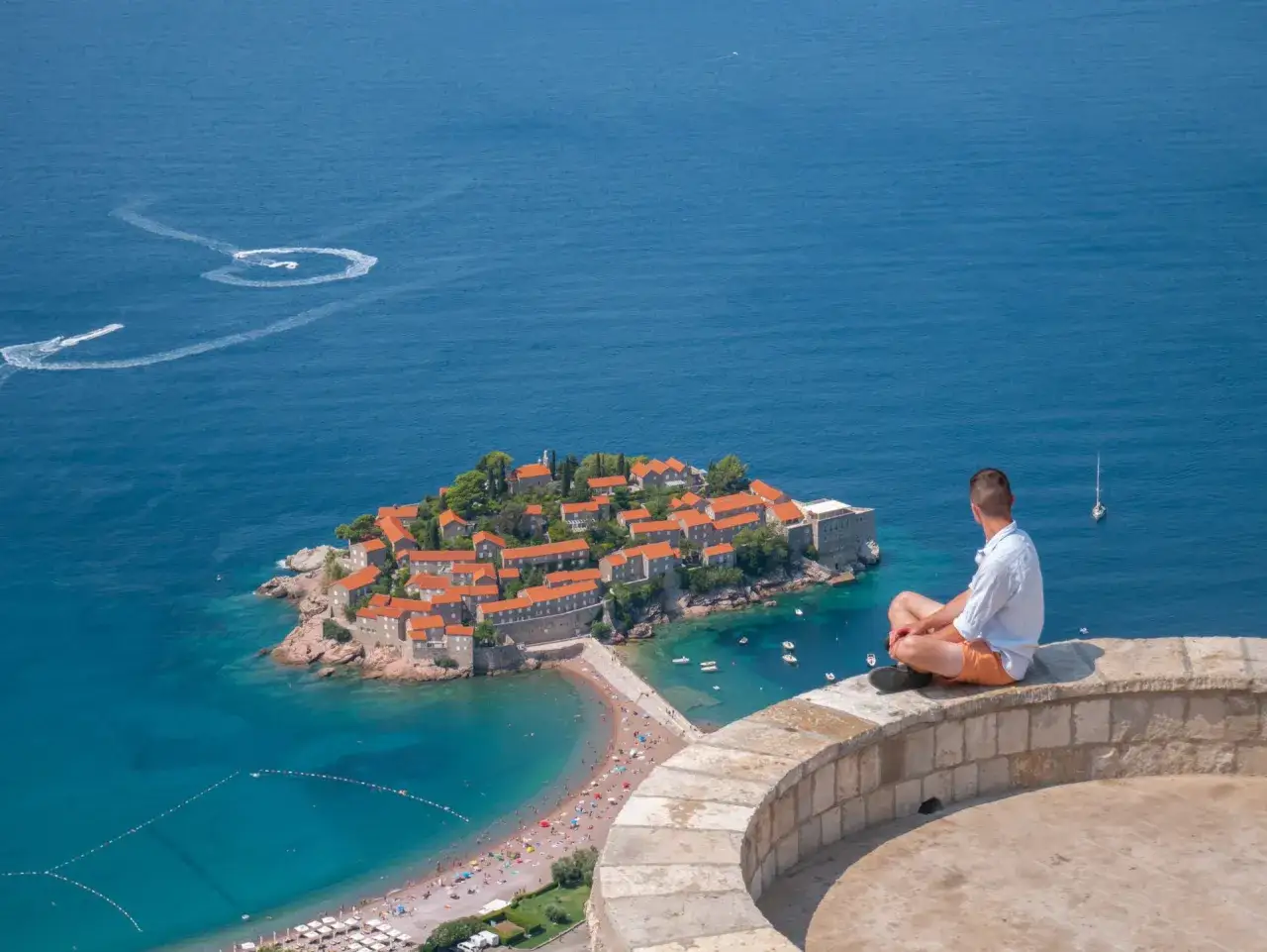 Czarnog&oacute;ra na co uważać? Widok na wyspę Sveti Stefan, turkusowe morze i plażę. Mężczyzna siedzi na murku, podziwiając krajobraz.
