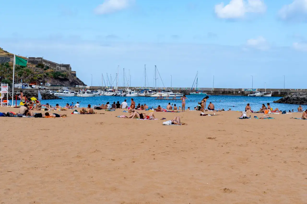 Plaża w Machico na Maderze, ludzie opalają się na piasku, w tle port z łodziami i fortyfikacje.