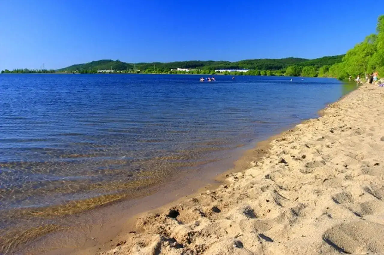 Słoneczny dzień nad zalewem Sulejowskim. Piaszczysta plaża, błękitna woda i zielone wzgórza w tle. Kilka osób pływa i odpoczywa.